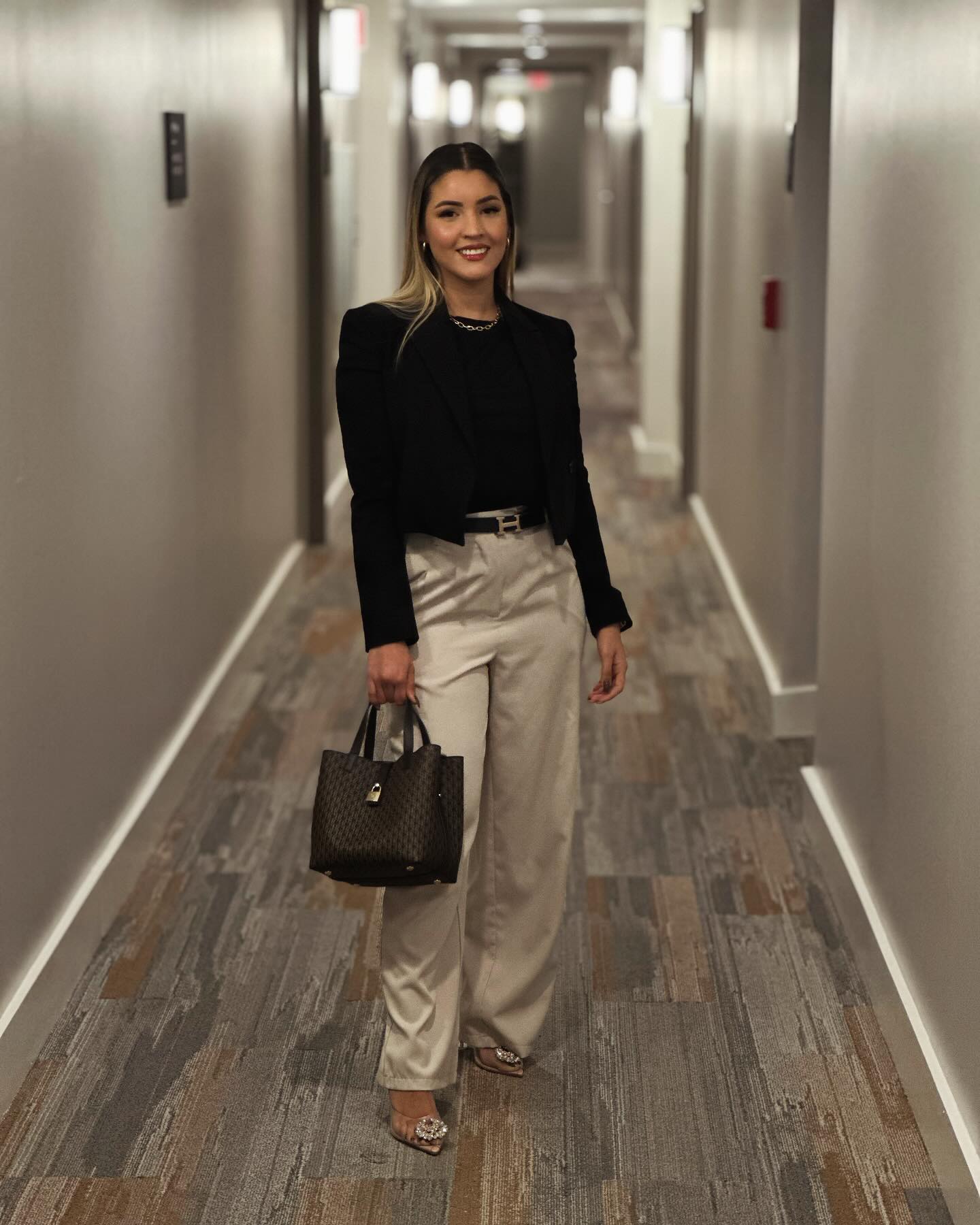 Luisa smiling in a black blazer, cream trousers and heels, holding a handbag in a hallway, with her whole outfit visible