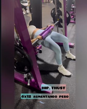 Frame from a gym reel showing Luisa performing a hip thrust exercise on purple gym equipment, wearing pale-blue leggings and white sneakers