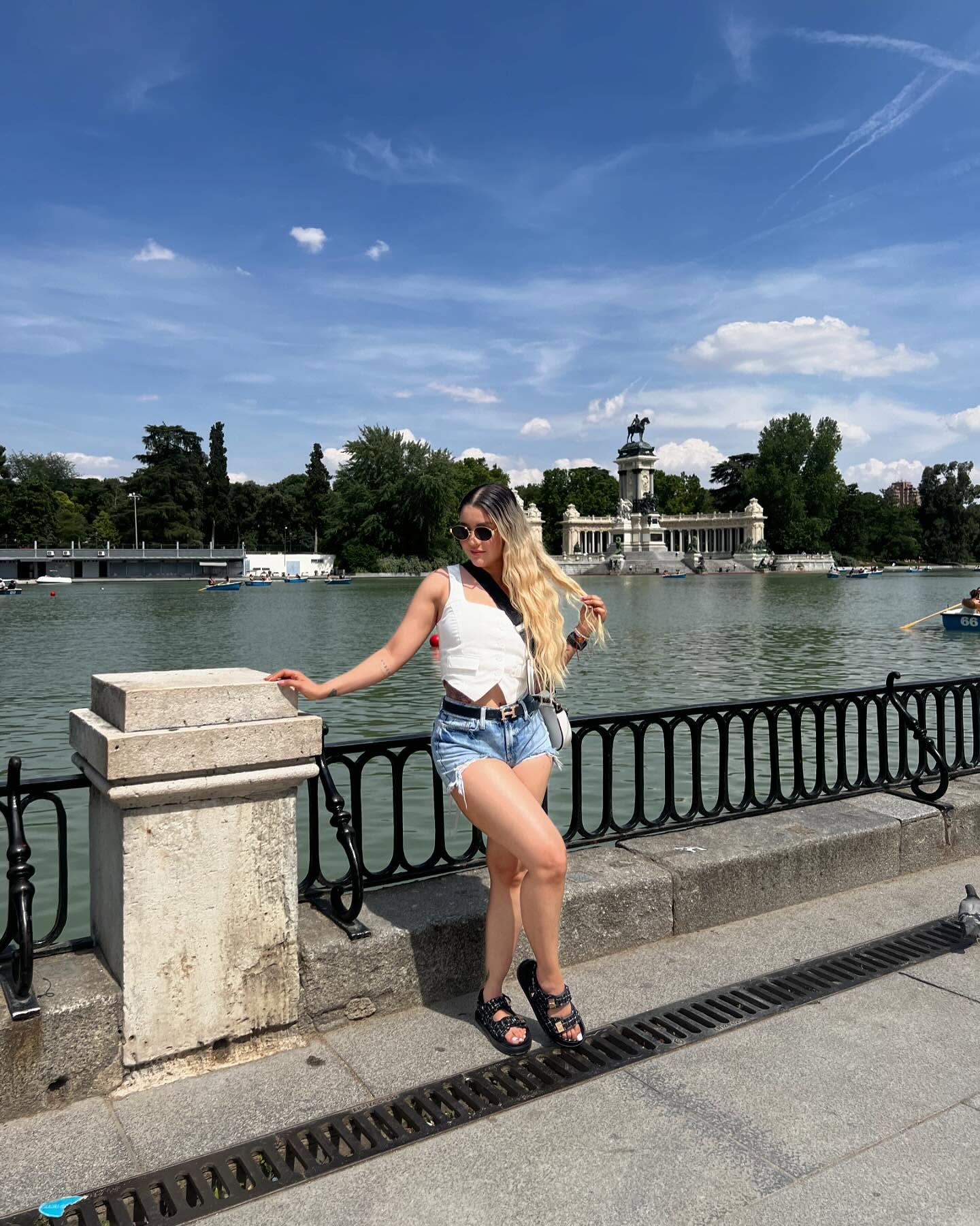 Luisa wearing a white top, denim shorts, sunglasses and black sandals beside a lake in Madrid