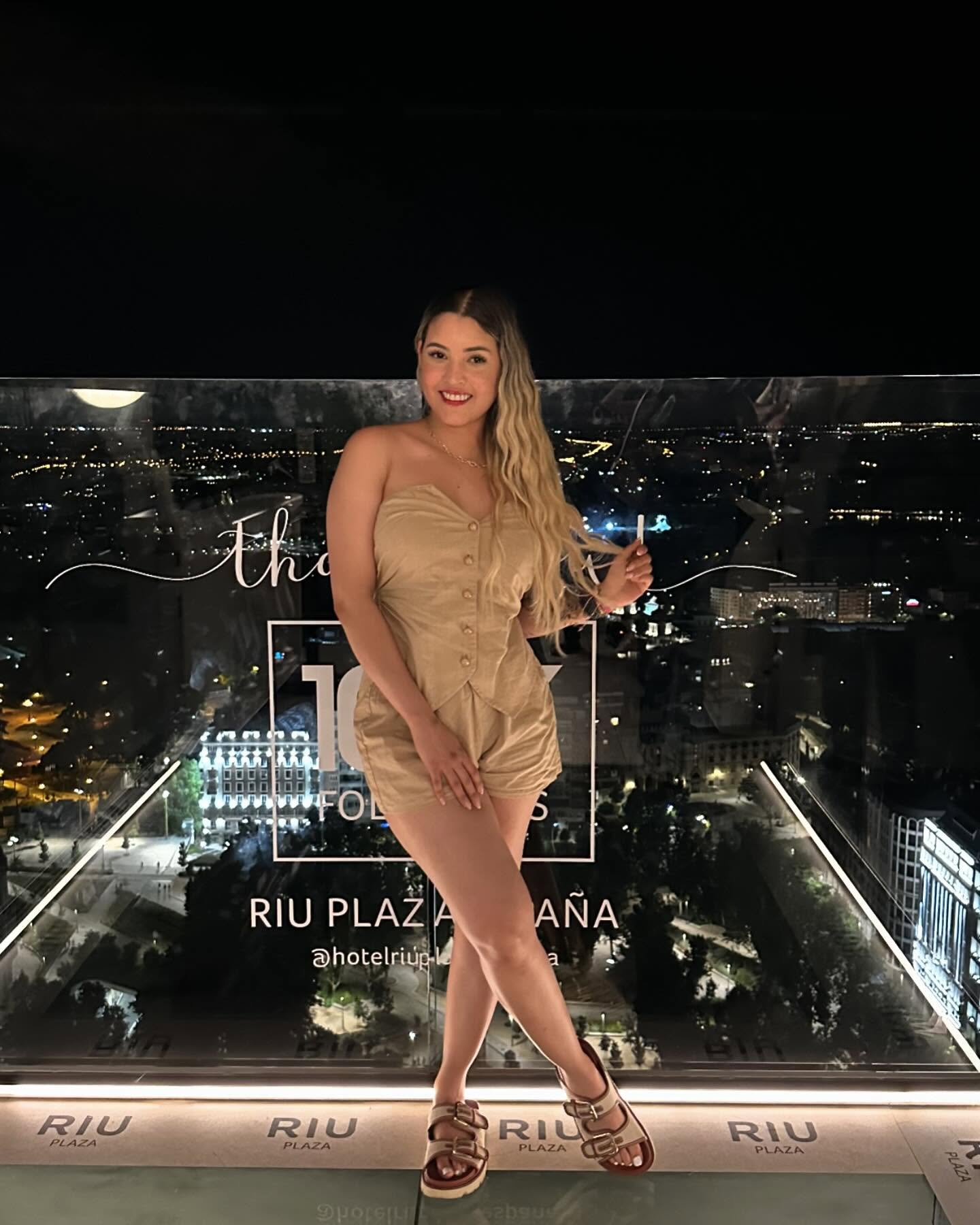Luisa smiling in a tan romper and sandals on a rooftop terrace overlooking Madrid at night