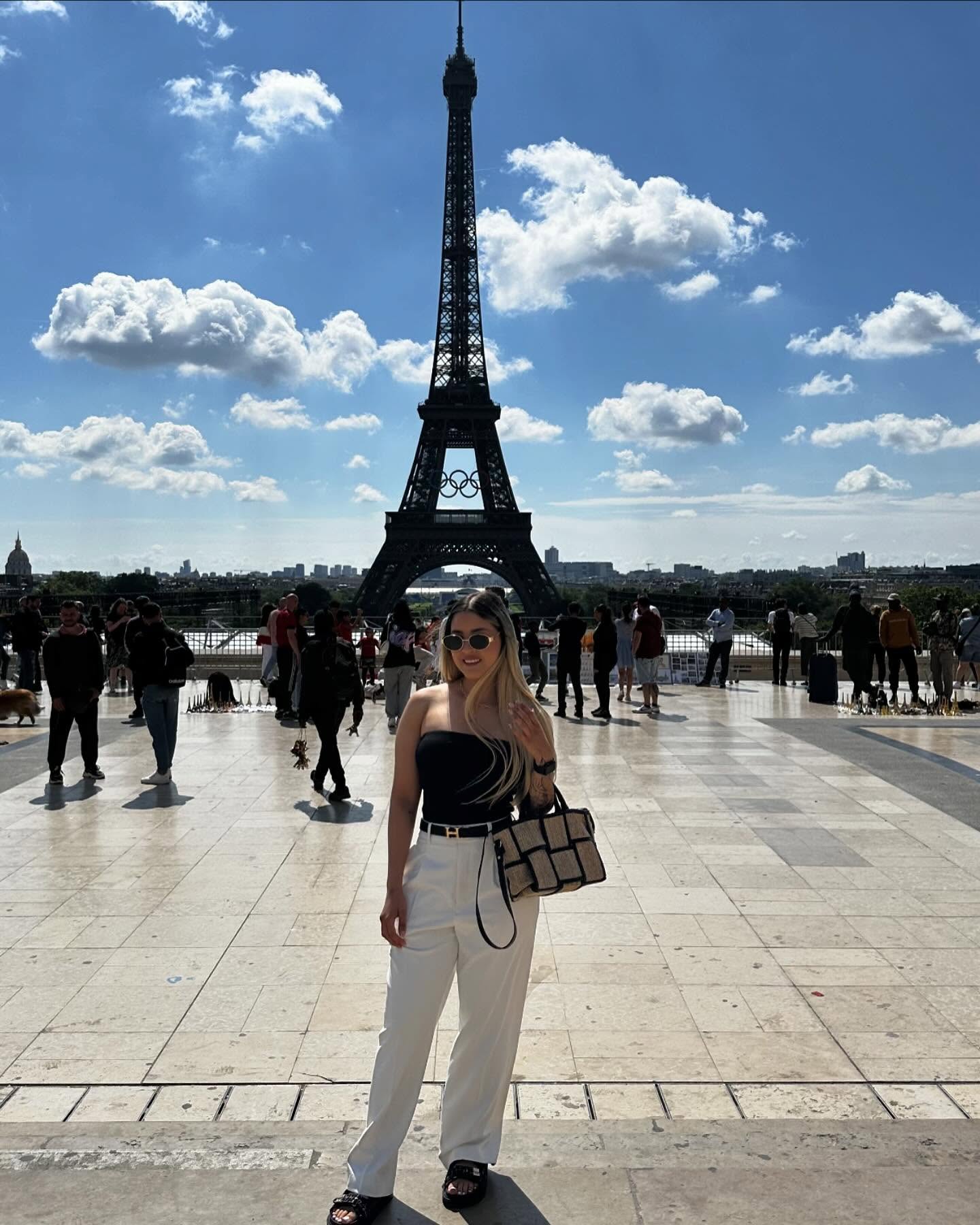 Luisa smiling in sunglasses, a black top and white trousers in front of the Eiffel Tower, with her whole outfit and sandals visible